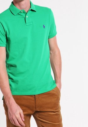 Man draagt een felgroen poloshirt met korte mouwen en een klein paars logo, evenals een bruine corduroy broek, poseert met één hand in de zak.