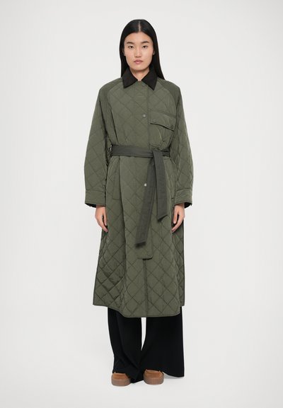 Cappotto verde oliva trapuntato con vita accentuata da una cintura, colletto e una singola tasca sul petto, indossato su pantaloni neri a gamba larga.