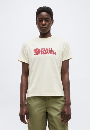 Άτομο που φοράει λευκό T-shirt με κόκκινο λογότυπο "Fjällräven" και χακί παντελόνι, στέκεται μπροστά σε απλό λευκό φόντο.