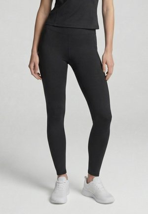 Woman wearing black leggings and white sneakers, standing against a plain light background with hands relaxed by sides.