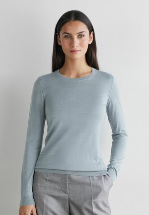 Kvinde med langt mørkt hår iført en lyseblå langærmet trøje og grå bukser, stående mod en ensfarvet lys væg.