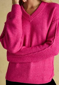 Pulover tricotat roz, cu decolteu în V, mâneci și tiv cu dungă. Materialul pare moale, cu o textură plăcută.