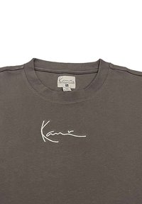 T-shirt en coton gris avec un col rond et une signature brodée blanche sur la poitrine. Étiquette visible avec des informations de taille.