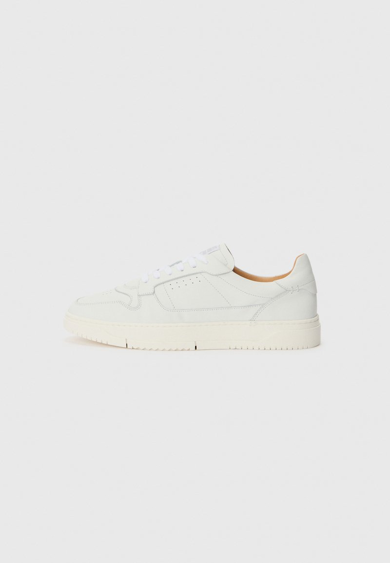 White leather sneaker with a textured surface, rounded toe, and perforated detailing. Features white laces and a light beige rubber sole.