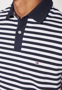 Polo tričko s horizontálními pruhy v námořnické a bílé barvě, s námořnickým límcem a malým vyšitým logem na hrudi.