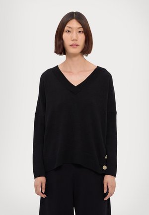Maglione nero lavorato a maglia con scollo a V, spalle scese e bordi a coste. Presenta bottoni decorativi dorati sull'orlo laterale. Texture liscia.