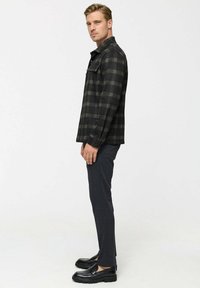 Schwarze karierte Shirtjacke mit zwei Brusttaschen und Kragen; kombiniert mit dunklen, schmalen Hosen und schwarzen Schuhen. Seitenansicht.