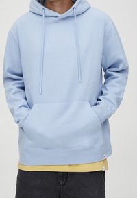 Personne portant un sweat à capuche bleu clair avec les mains dans la poche avant, superposé à un t-shirt jaune et assorti d'un pantalon foncé.