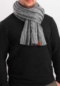 Knitted gray scarf with a textured pattern, featuring a leather tag. Worn around the neck over a black sweater.