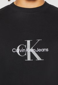 Černé bavlněné tričko s stříbrně vyšitým logem "CK" a textem "Calvin Klein Jeans" pod ním. Klasický design s kulatým výstřihem.