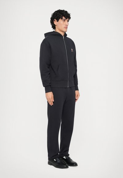 Sudadera con cremallera de color navy, hecha de algodón, con un bolsillo tipo canguro, combinada con pantalones jogger a juego y zapatos negros. Presenta un pequeño logo en el pecho.