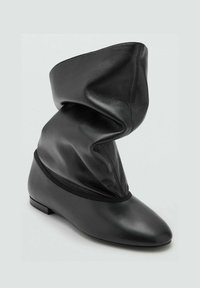 Bota de cuero negro con un diseño holgado en la parte superior, punta afilada y suela plana. El diseño presenta una textura suave y arrugada, además de una banda elástica en el tobillo.