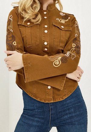 Person trägt eine braune, geknöpfte Jacke mit bestickten Blumenmustern an den Ärmeln und blaue Jeans, die Arme vor dem Oberkörper verschränkt.