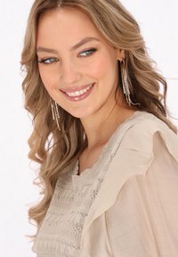 Beige Bluse mit strukturiertem Strickmuster, Rüschensleeves und weichem Stoff. Lange, funkelnde Ohrringe. Leichtes Make-up und lockiges Haar.