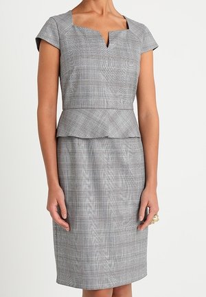 Femme portant une robe peplum à carreaux gris avec des manches courtes et un décolleté carré en encoche, debout devant un fond blanc uni.