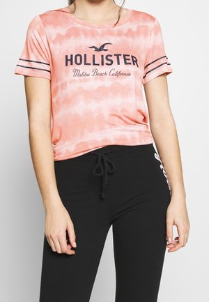 T-shirt rose à manches courtes en tie-dye avec le graphique "Hollister" en noir. Col rond et accents de manches rayées sur un pantalon noir ajusté.