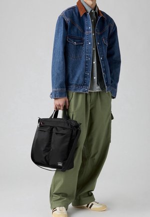Homme portant une veste en denim, une chemise à carreaux, un pantalon cargo olive, des baskets beiges, tenant un sac cabas noir avec plusieurs poches.