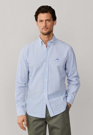 Homme portant une chemise à rayures verticales bleues et blanches avec col et boutons, debout avec une main dans la poche sur un fond uni.