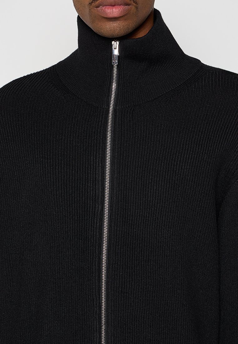 Schwarzer Strickpullover mit hohem Kragen und silbernem Reißverschluss, der über eine gerippte Textur und eine taillierte Passform verfügt.