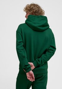 Zielona bluza z kapturem wykonana z miękkiego materiału, z dużym kapturem i luźnym krojem. Widoczne ściągacze przy rękawach i u dołu, o gładkiej fakturze.