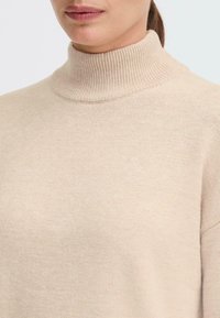 Bežs augstais apkakles džemperis mīkstajā dzijas audumā, ar ribotu apkakli un brīvu piegriezumu. Gluda tekstūra ar minimālu dizainu.