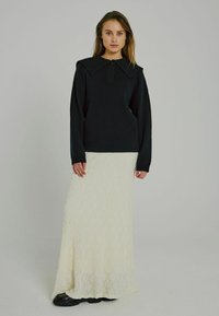 Schwarzer Strickpullover mit großem Kragen und vorderem Knopf, kombiniert mit einem langen, cremefarbenen, strukturierten Maxiröck. Schwarze Schuhe werden getragen.