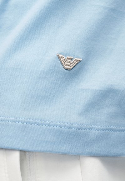 Λιλά ύφασμα με μικρό κεντημένο ασημένιο λογότυπο αετού πάνω από ραμμένη γαζωτή και λευκό ύφασμα από κάτω.