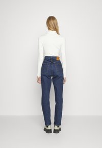 Jeans de mezclilla azules de cintura alta con bolsillos traseros y una etiqueta de cuero. Llevados con un ajustado cuello de tortuga blanco de canalé y zapatos negros.
