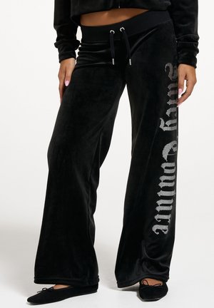Schwarze Velours-Hose mit weitem Bein, Kordelzugbund und funkelndem silbernem Schriftzug an einem Bein, getragen mit schwarzen Ballettschuhen.