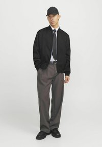 Chaqueta de bombardero negra, camisa blanca y corbata negra, combinadas con pantalones grises y zapatos negros. El modelo lleva una gorra negra a juego.