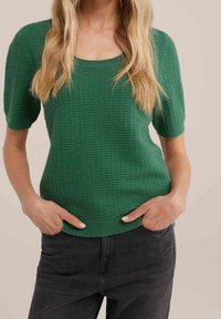 Haut tricoté texturé vert avec un décolleté carré et des manches courtes, associé à un jean foncé. Le tissu présente un motif côtelé.