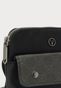 Črna tkaninska torbica s srebrnim zadrgo, sprednjim žepnim pokrovom s teksturiranim vzorcem in zatičnim gumbom ter okroglim kovinskim logotipom "V".