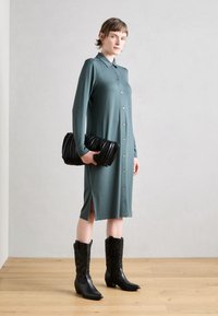 Robe longueur genou, à boutons, en tissu doux bleu-vert, avec manches longues et fentes latérales, associée à une pochette texturée noire et des bottes de cowboy.