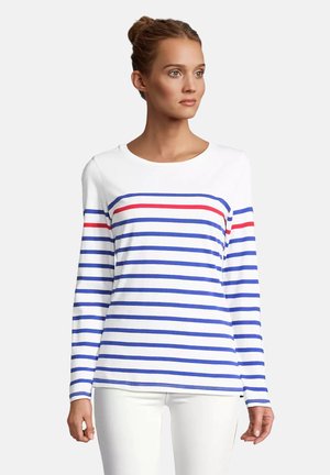 Donna che indossa un top bianco a maniche lunghe con strisce orizzontali blu e una rossa sul petto, abbinato a pantaloni bianchi, guarda verso destra.