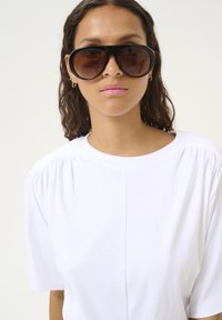 Zwarte oversized zonnebril met een verlooplens in bruin, gedragen met een wit shirt met korte mouwen dat zachte plooien bij de schouders heeft.