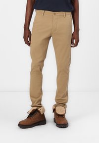 Man wearing slim-fit beige chinos and brown lace-up boots with fleece-lined cuffs against white background.