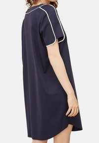 Donna che indossa un abito corto blu navy a maniche corte, largo, con profili bianchi sulle maniche e sul collo, in piedi davanti a uno sfondo neutro.