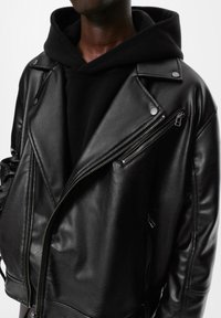 Crna kožna motorkarska jakna s asimetričnim patentnim zatvaračem, srebrnim detaljima i podstavljenim ovratnikom, složena preko crne dukserice s kapuljačom.