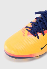 Jasne žlté futbalové topánky s textúrovaným zvrškom, námorníckymi šnúrkami a červenými akcentmi. Obsahujú výrazné predlžené logo Nike a tvarované kolíky.