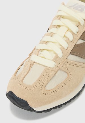 Gros plan d'une basket beige et crème avec des panneaux en suède, des lacets crème et un embout et une semelle en caoutchouc noir.
