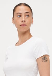 Biele rebrované tričko s krátkymi volánovými rukávmi. Má okrúhly výstrih. Modelka má na ľavom ramene tetovanie a malé kruhové náušnice.