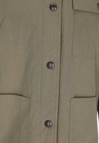 Cămașă khaki cu închidere pe butoane, două buzunare pe piept și butoane metalice rotunde. Materialul este neted, cu un ușor luciu.