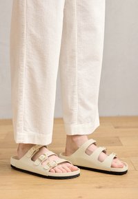 Lyse beige sandaler med tre justerbare stropper med metallspenner, en polstret fotbed og såle i svart gummi.