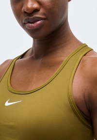 Zbliżenie osoby noszącej oliwkowo zielony sportowy top Nike, na którym widoczne są dekolt, ramię oraz biały logo Nike na klatce piersiowej.