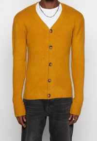Hořčicově žlutý cardigan z měkké vlny s výstřihem do V, pěti hnědými knoflíky a žebrovanými manžetami. Nosený přes bílou košili.