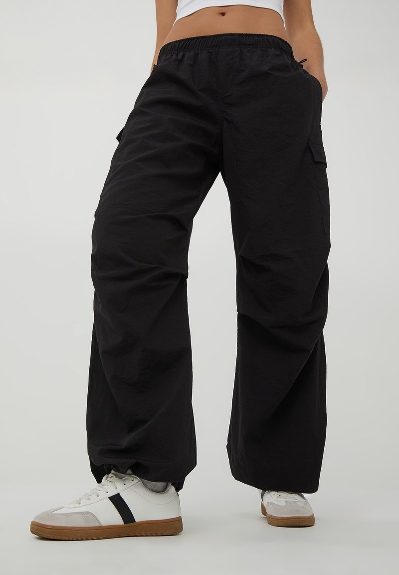 Personne portant un pantalon cargo noir ample avec une ceinture élastique et des baskets blanches avec une bande noire, debout devant un fond uni.