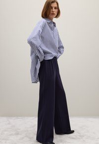 Gestreepte blauw-witte shirt met een kraag en zijsplitten, gecombineerd met wijde marineblauwe broek. Soepele textuur, ontspannen ontwerp, minimale accenten.