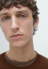 Přiblížení mužského obličeje se středně dlouhými kudrnatými hnědými vlasy, zelenomodrýma očima, světlou pokožkou a jemnými kožními nedokonalostmi. Žádné viditelné doplňky.