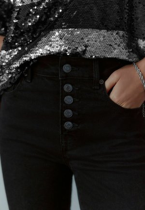 Schwarze hochtaillierte Jeans mit fünf schwarzen Knöpfen an der Vorderseite, kombiniert mit einem schwarzen Pailletten-Top, das eine glänzende, strukturierte Oberfläche zeigt.
