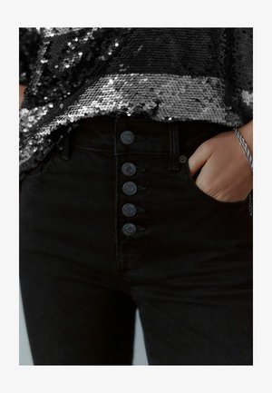 Jeans neri a vita alta con cinque bottoni neri davanti, abbinati a un top di paillettes nere che mostra una superficie lucida e texturizzata.